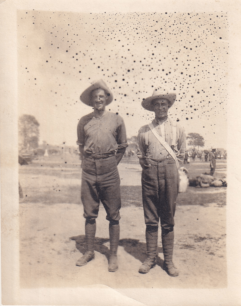 Two soldiers standing together in a field, wearing military uniforms and wide-brimmed hats, with a crowd in the background.